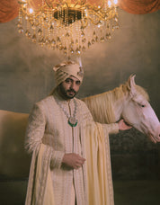 A man in an ornate cream and gold embroidered sherwani with a matching beige turban stands beside a white horse under a large crystal chandelier.