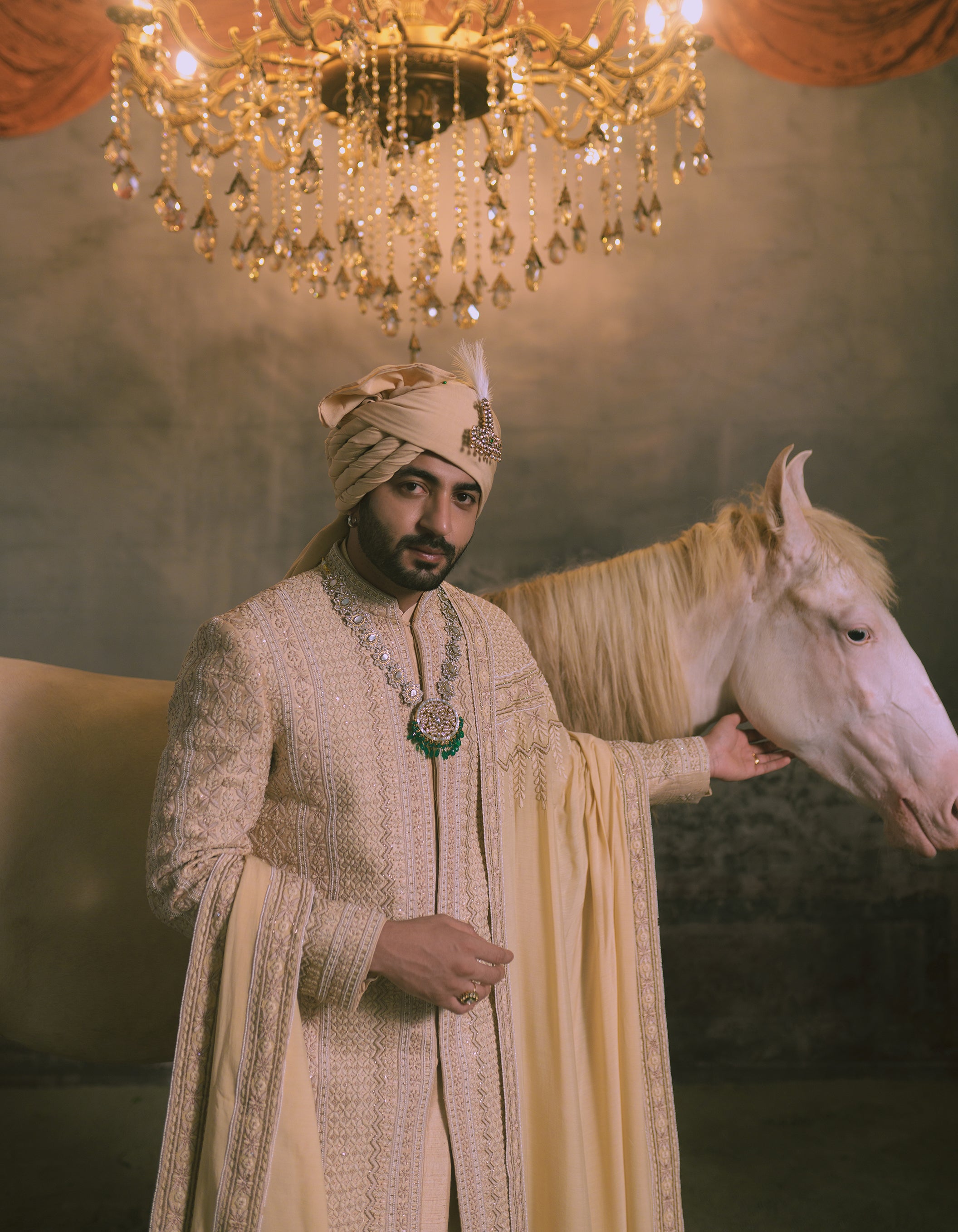 A man in an ornate cream and gold embroidered sherwani with a matching beige turban stands beside a white horse under a large crystal chandelier.