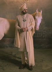 A man in an ornate golden sherwani and matching turban stands beside a white horse in a dramatic studio setting with draped orange fabric, a large chandelier, and a textured gray backdrop.