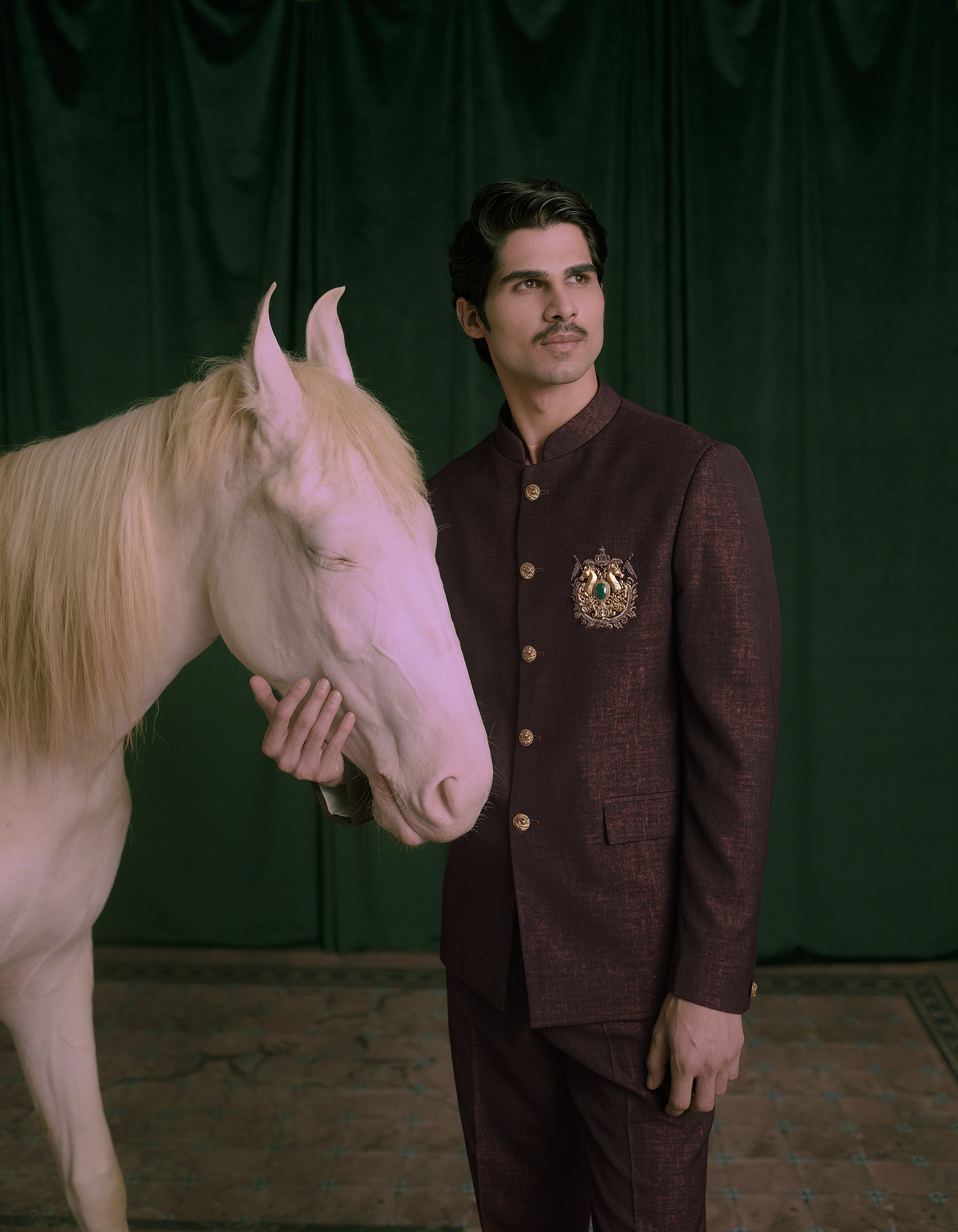 A man in a dark maroon textured jodhpuri/bandhgala suit with gold buttons and a crest on the chest.